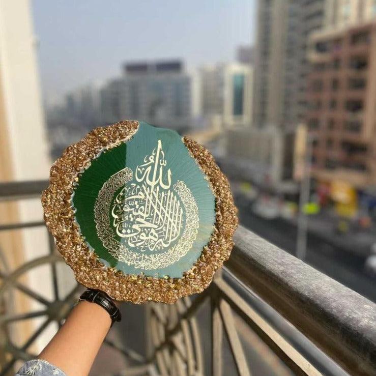 "Round decorative Islamic display plate featuring the sacred Ayatul Kursi verse in white calligraphy on a green background, surrounded by a gold textured border, held outdoors with a city skyline in the background."