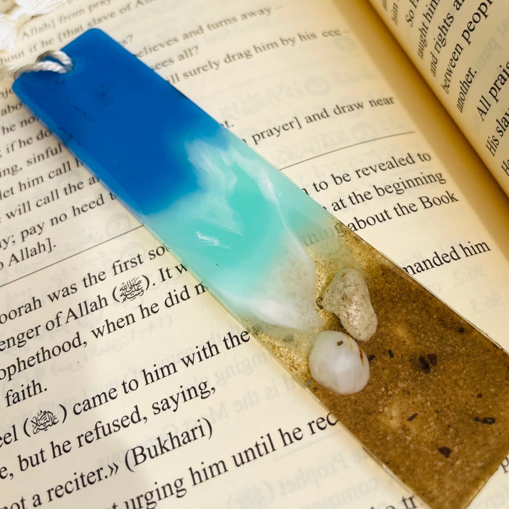 Resin bookmark with an ocean theme placed on an open book, featuring blue hues, wave patterns, and a glossy epoxy finish, adding beauty to the reading experience.